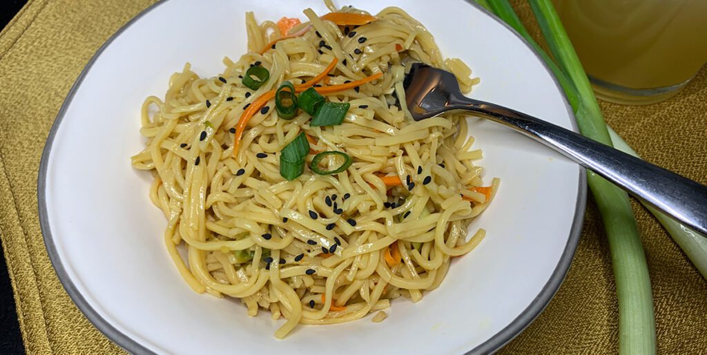 sesame noodles in a white bowl with a fork showing noodles, cucumbers, carrots and sesame seeds.