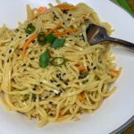 sesame noodles in a white bowl with a fork showing noodles, cucumbers, carrots and sesame seeds.