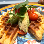 Plated savory waffles with avocado, honey, tomatoes, greens and sprinkled with salt and pepper